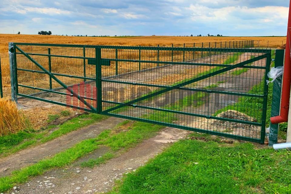 Zielona brama wjazdowa z siatki na tle pola zboża, z widocznymi śladami opon na trawiastym podłożu i częściowo owiniętym słupkiem po prawej stronie.