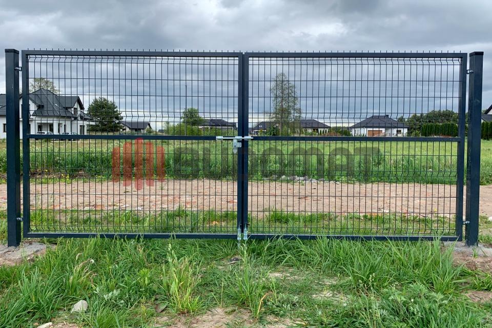 Dwuskrzydłowa brama z paneli zgrzewanych, zabezpieczona zamkiem na tle domów jednorodzinnych i pochmurnego nieba.