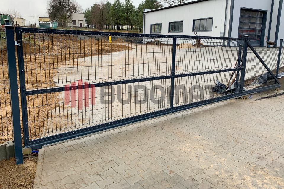 Przesuwna brama wjazdowa z paneli metalowych, widoczna na tle budynku przemysłowego z placem wyłożonym kostką brukową.