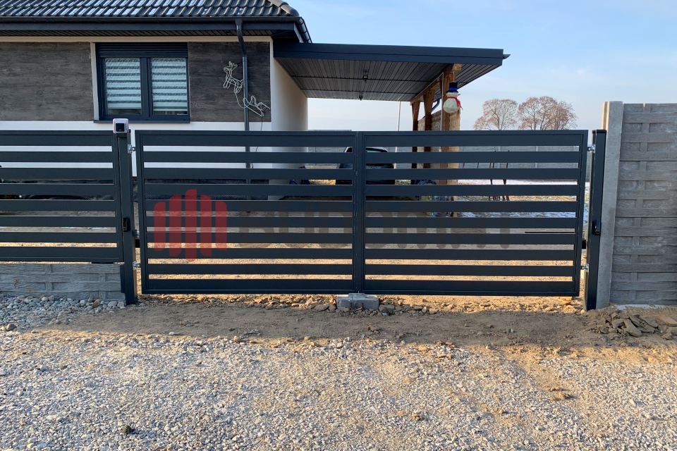 Nowoczesna, pozioma brama wjazdowa z metalowymi przęsłami w kolorze antracytowym, osadzona pomiędzy betonowymi słupkami, na tle domu z dekoracją świąteczną i zadaszeniem garażowym.