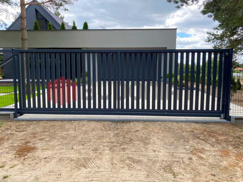 Nowoczesna, przesuwna brama wjazdowa z metalowymi sztachetami w kolorze grafitowym, zamontowana na betonowej podmurówce, z widocznym fragmentem ogrodzenia z siatki w tle i nowoczesnym domem...