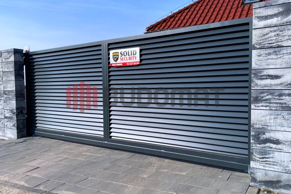 Przesuwna, pozioma brama metalowa w kolorze grafitowym, z tabliczką firmy ochroniarskiej Solid Security, zamontowana między dwoma słupkami z szarego betonu architektonicznego, na tle kostki...