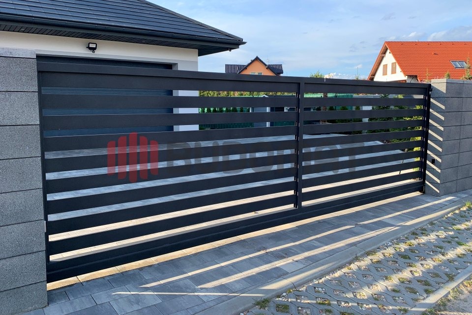 Nowoczesna, przesuwna brama wjazdowa z poziomych, czarnych profili, osadzona między szarymi słupkami z bloczków betonowych, na tle kostki brukowej i domów.