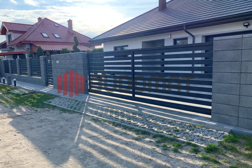 Nowoczesna, pozioma brama wjazdowa z szarymi, betonowymi słupkami i przęsłami ogrodzenia, uzupełniona kostką ażurową na podjeździe, w tle dom z czerwonym dachem.