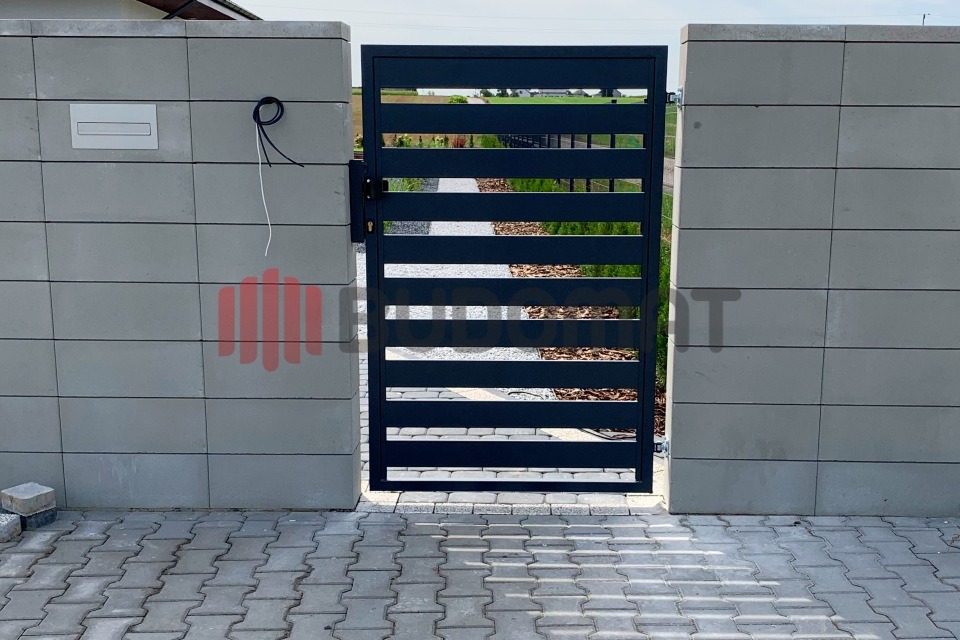 Nowoczesna, pozioma brama metalowa w kolorze antracytowym, zamontowana między betonowymi słupkami z widoczną skrzynką na listy i wystającymi przewodami, na tle brukowanej nawierzchni i zieleni.