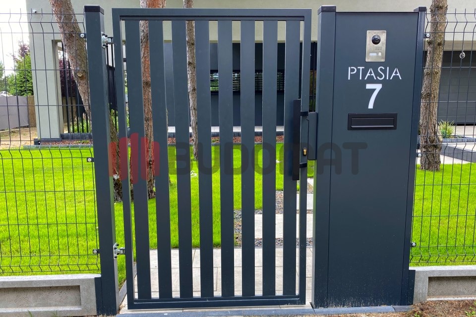 Nowoczesna, grafitowa furtka z pionowych profili, wkomponowana w siatkę ogrodzeniową i betonową podmurówkę, z numerem domu 'Ptasia 7' i wideodomofonem.