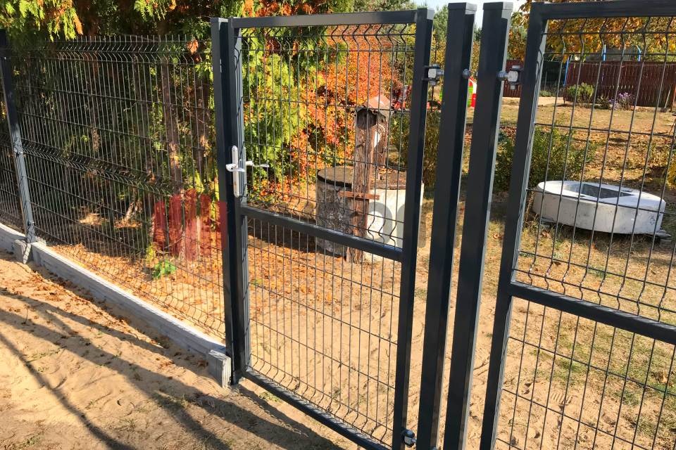 Zamknięta furtka w systemie ogrodzeniowym z paneli zgrzewanych, szara kolorystyka, widoczne betonowe podmurówki i słupki, w tle drzewa z jesiennymi liśćmi.