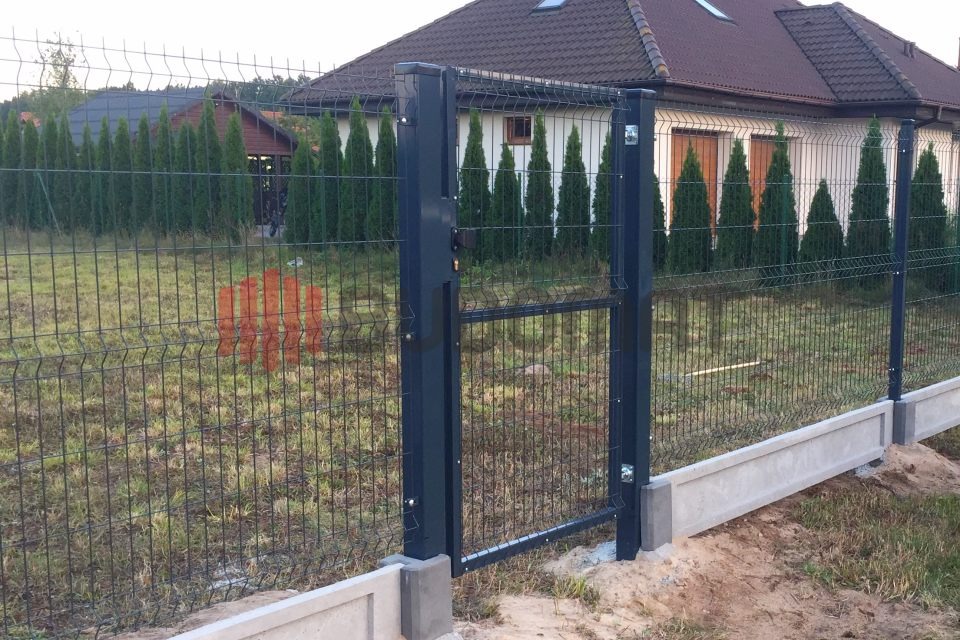 Ciemnoszara, metalowa brama wjazdowa z siatki panelowej na betonowej podmurówce, widok z zewnątrz posesji, w tle dom jednorodzinny i rząd tuj.