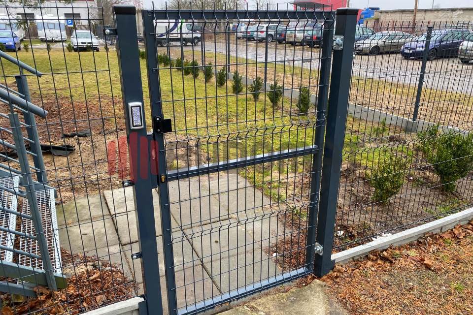 Szara, metalowa furtka z panelem numerycznym na słupku, część ogrodzenia panelowego, z widocznymi młodymi krzewami wewnątrz posesji i parkingiem w tle.