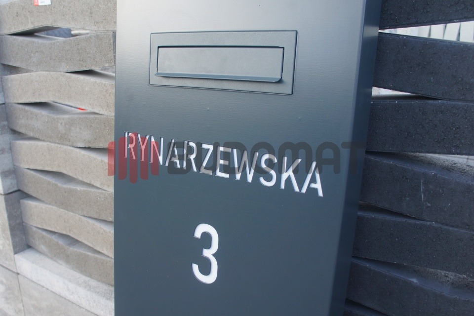 Szary słupek z numerem domu i nazwą ulicy, stanowiący część ogrodzenia z betonowych elementów o nieregularnej fakturze.