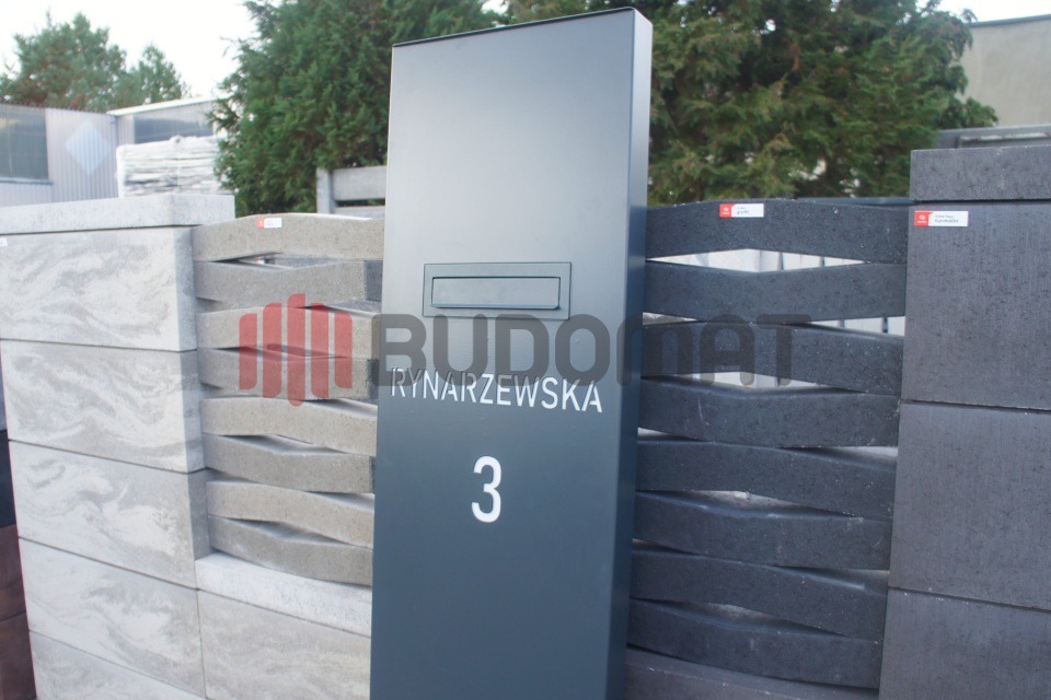 Fragment ekspozycji różnych wariantów kolorystycznych i wzorów bloczków do budowy ogrodzeń modułowych z betonu architektonicznego, z szarą skrzynką na listy z numerem domu.