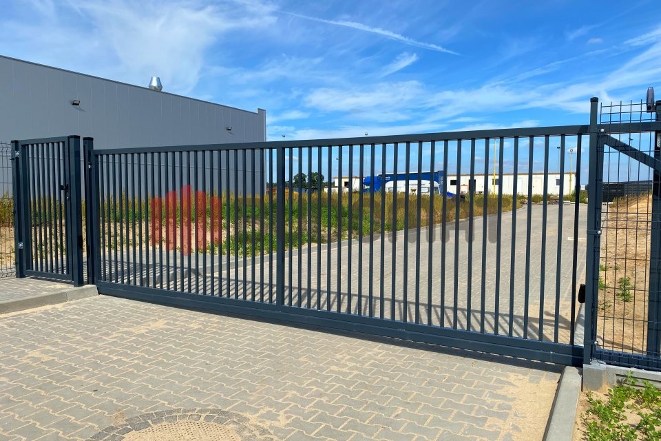 Przesuwna brama wjazdowa z metalowymi prętami, widoczna z zewnątrz posesji, z budynkiem przemysłowym w tle i kostką brukową na pierwszym planie.