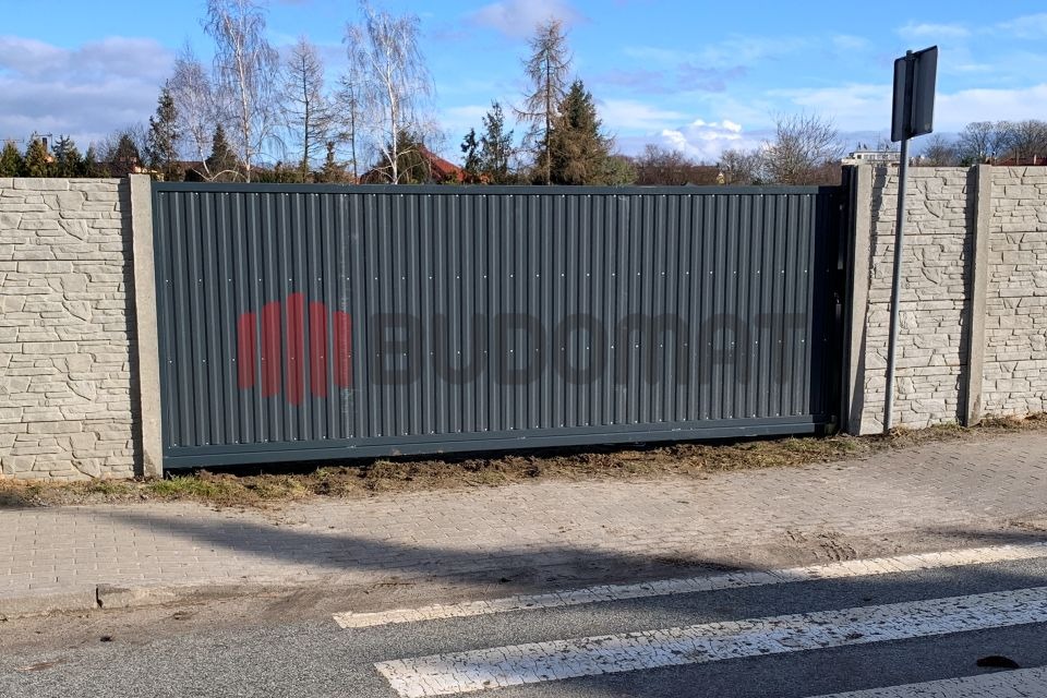 Szara, przesuwna brama wjazdowa z pionowymi panelami, zamontowana między dwoma słupkami z imitacji kamienia, widok z ulicy na tle drzew.