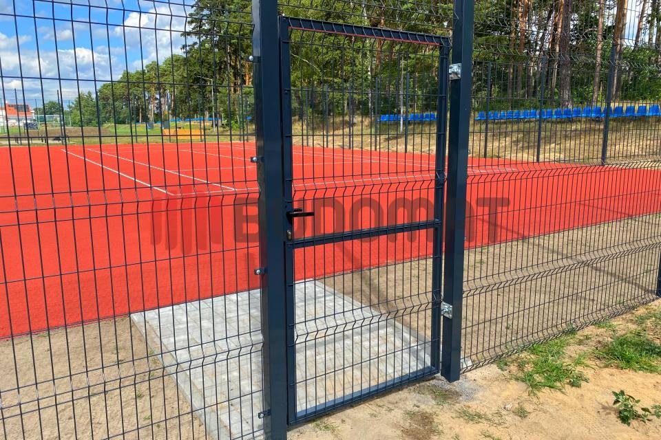 Ciemnoszara metalowa furtka z siatki ogrodzeniowej prowadząca na czerwone boisko sportowe, widok z zewnątrz.