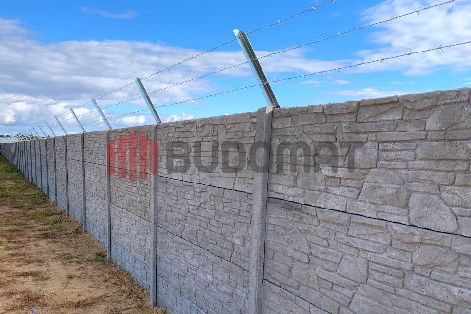 Długi, szary mur z prefabrykatów betonowych imitujących kamień, zwieńczony drutem kolczastym, ciągnący się wzdłuż piaszczystej ścieżki pod błękitnym niebem z chmurami.