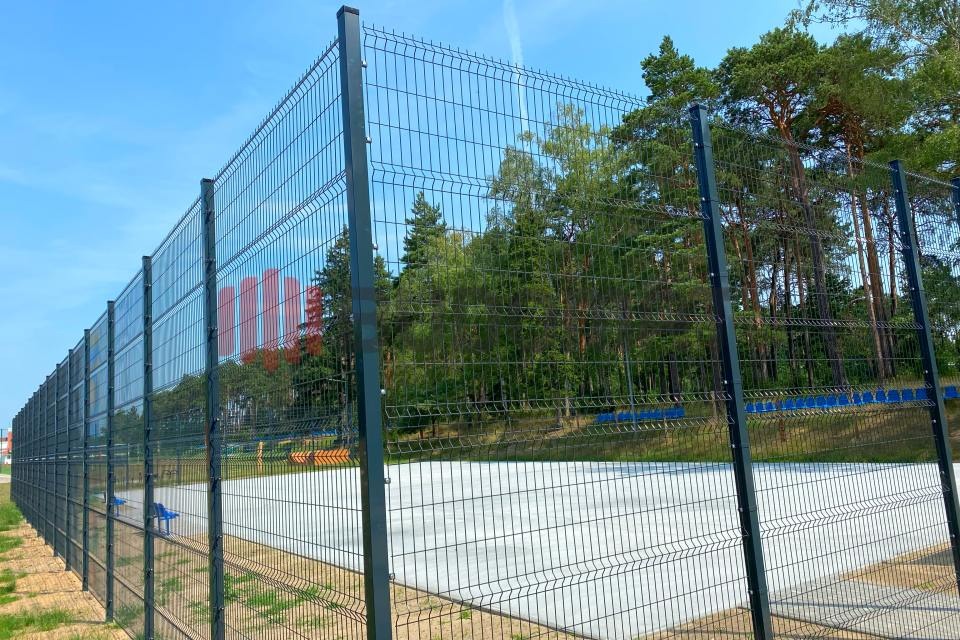 Długie, ciemne ogrodzenie panelowe z siatki zgrzewanej, otaczające betonowy plac zabaw, z widocznymi drzewami w tle i błękitnym niebem.