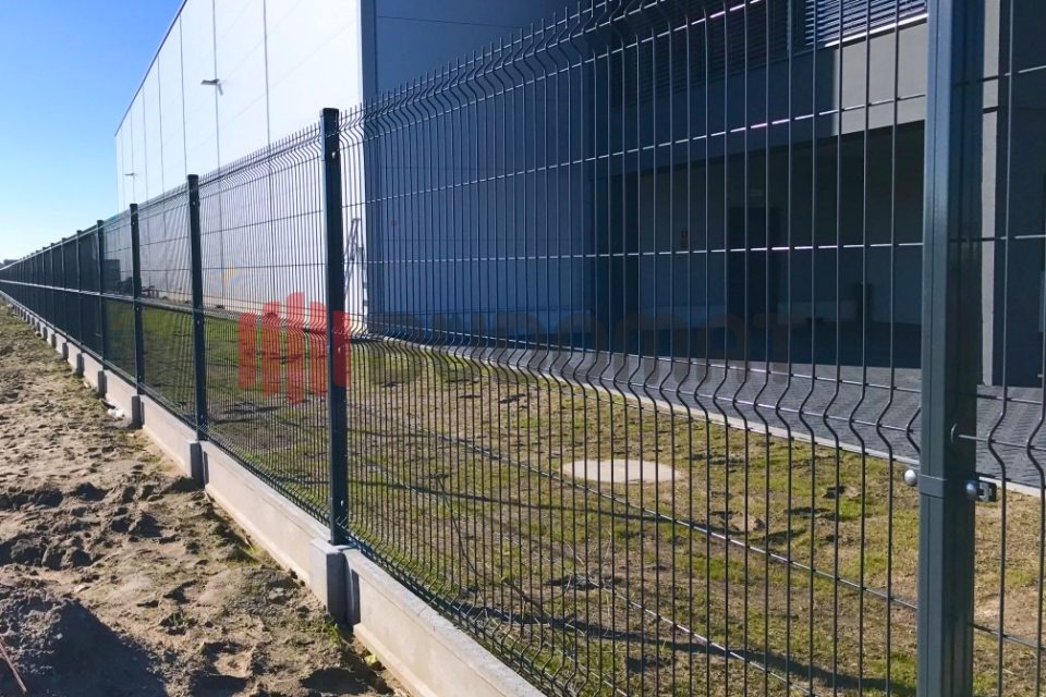 Długi, ciemnoszary panelowy system ogrodzeniowy z podmurówką betonową, zabezpieczający teren przemysłowy z widocznym budynkiem w tle.