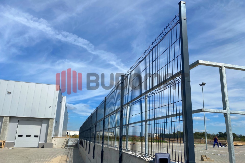 Długa, prosta linia ogrodzenia panelowego z metalowych prętów w kolorze grafitowym, oddzielająca teren przemysłowy z budynkami magazynowymi od parkingu, widok pod kątem w słoneczny dzień.