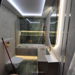 Remonty i Wykończenia Grzegorz Gołębiowski - Nowoczesna łazienka z podświetleniem LED, marmurowymi płytkami, złotą armaturą, owalnym lustrem z oświetleniem oraz toaletą przykrytą folią ochronną.