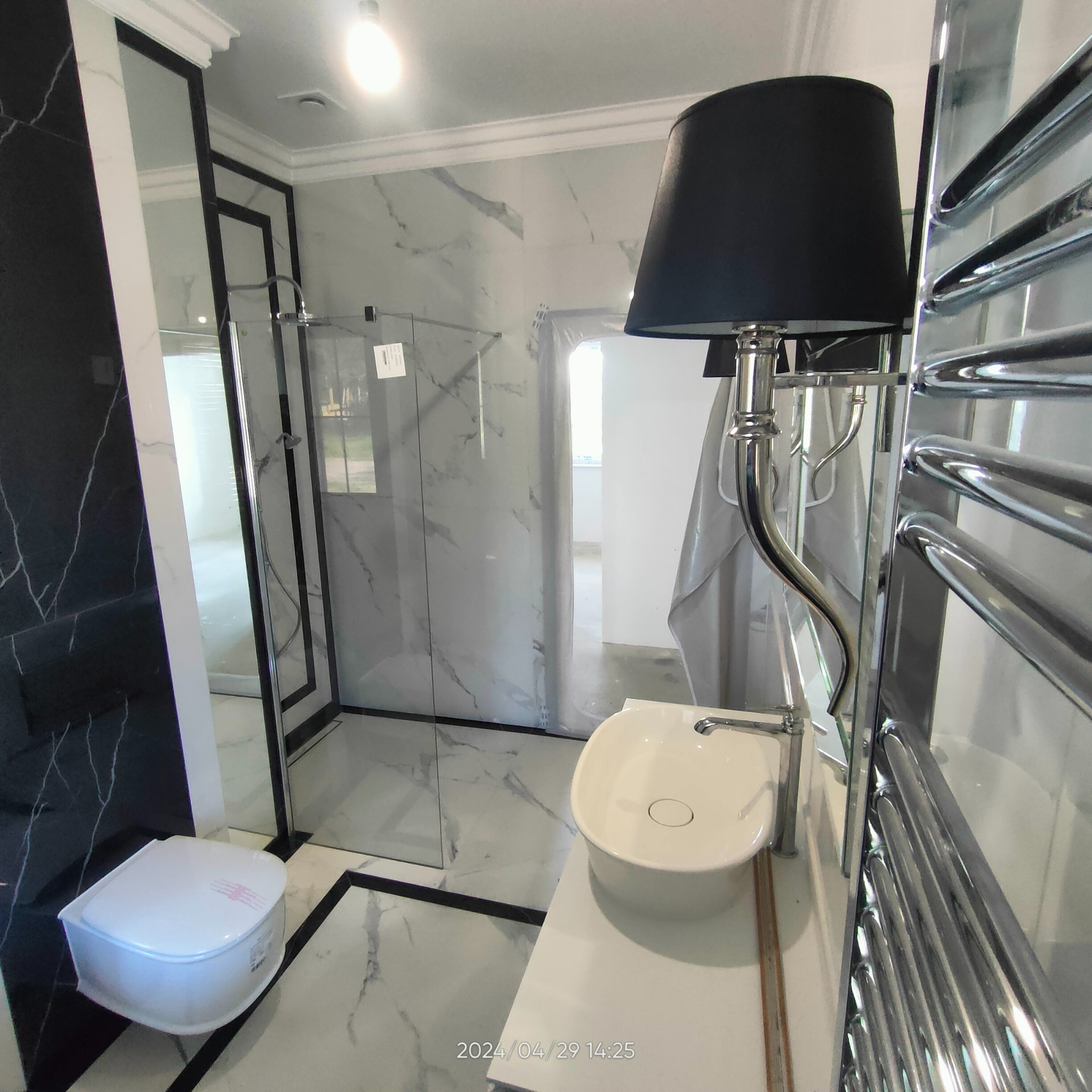 Nowoczesna łazienka z kabiną prysznicową typu walk-in, umywalką nablatową i grzejnikiem drabinkowym, wykończona płytkami imitującymi marmur z czarnymi akcentami, widoczna lampa stołowa z czarnym...