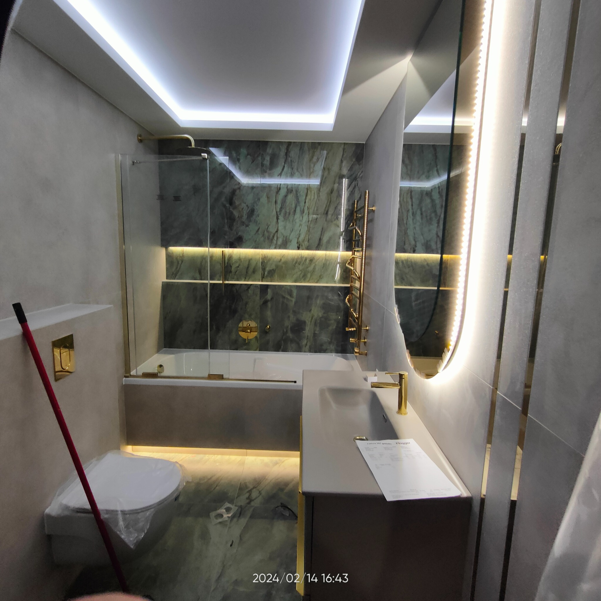 Nowoczesna łazienka z podświetleniem LED, marmurowymi płytkami, złotą armaturą, owalnym lustrem z oświetleniem oraz toaletą przykrytą folią ochronną.