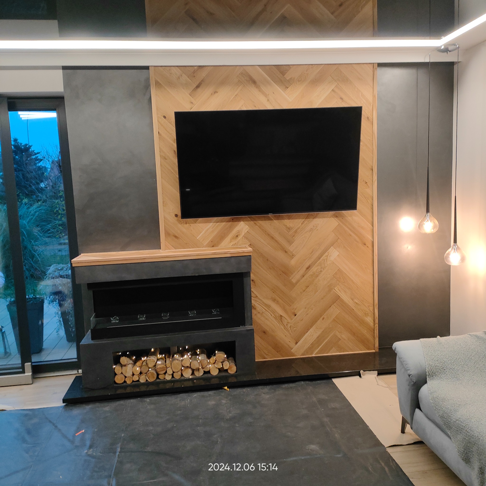 Salon w trakcie wykańczania: ściana z drewnianą okładziną w jodełkę, czarny kominek z drewnem, duży telewizor, szara ściana, lampy wiszące, podłoga zabezpieczona folią.