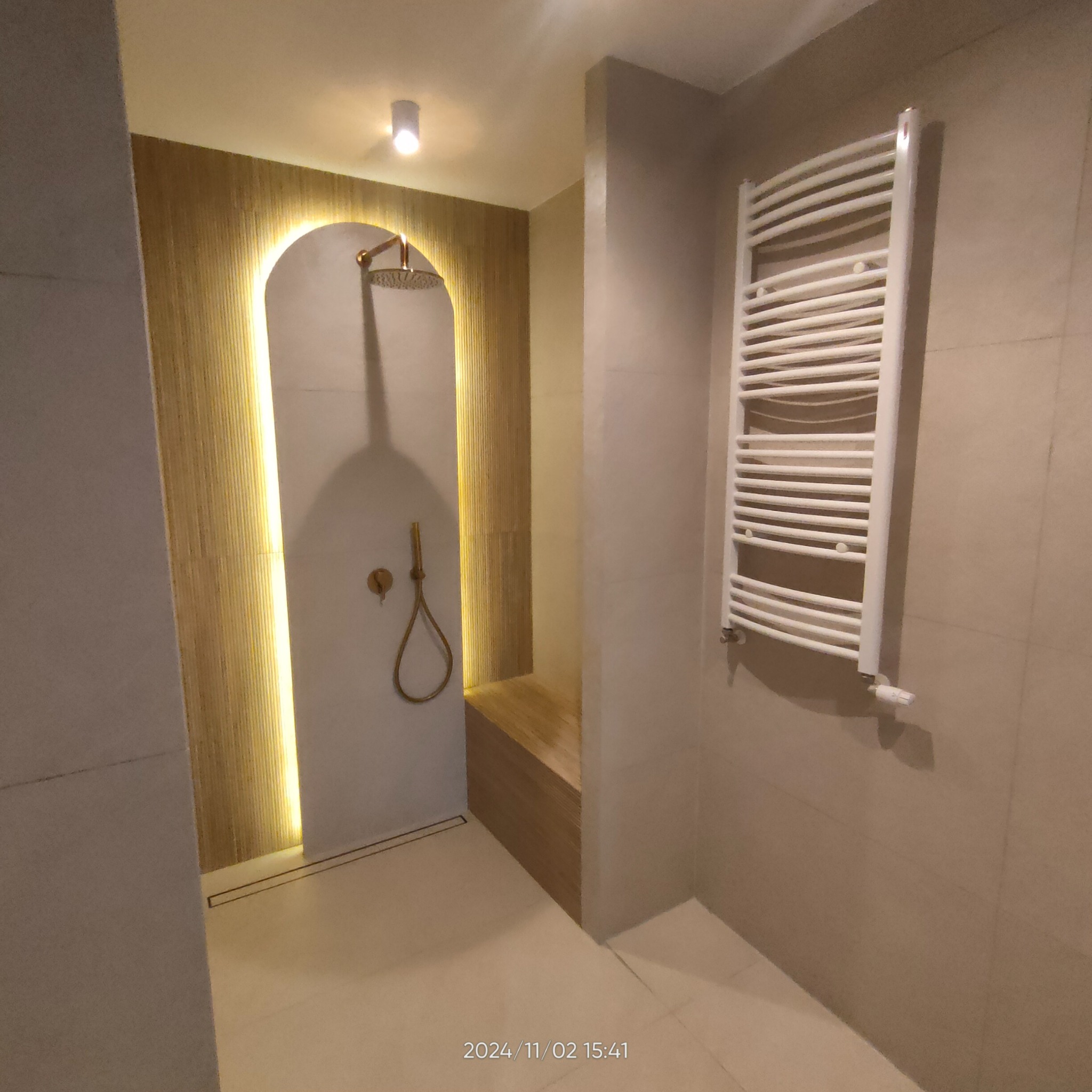 Nowoczesna łazienka z kabiną prysznicową typu walk-in, wykończoną płytkami w odcieniach beżu, z elementami dekoracyjnymi z drewna i oświetleniem LED. Widoczny grzejnik drabinkowy.