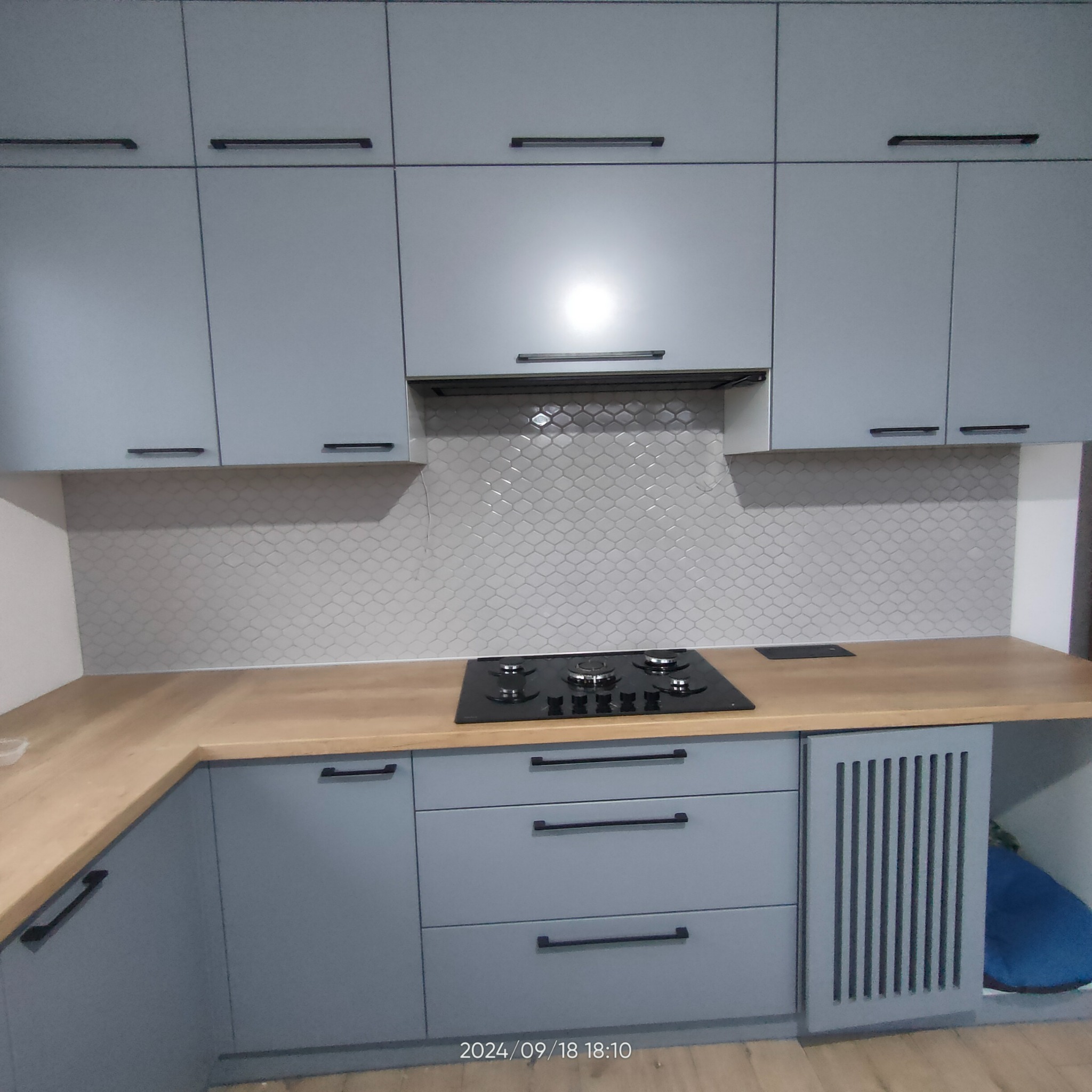 Szare szafki kuchenne z drewnianym blatem i czarną płytą grzewczą, tło z mozaiki w kształcie rybiej łuski, nowoczesne uchwyty.