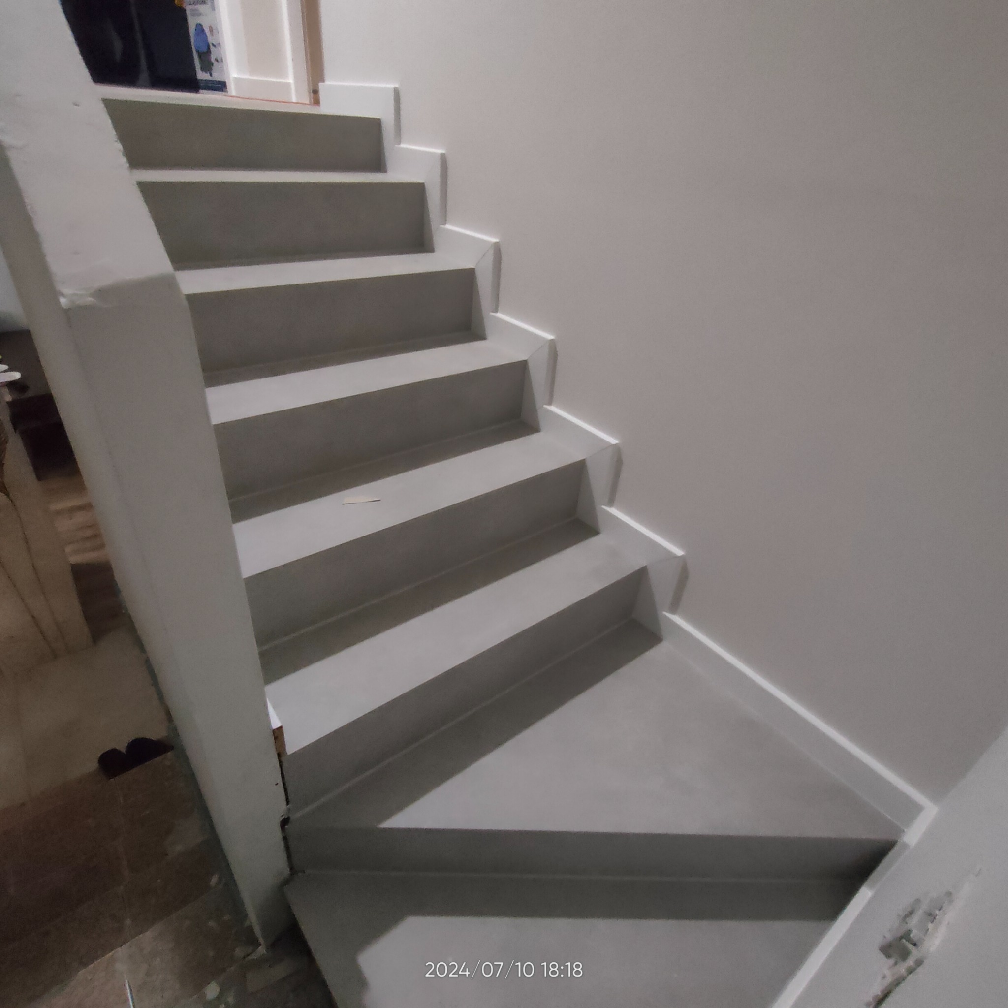 Szare schody wewnętrzne z białymi listwami przypodłogowymi, widok z dołu, ściana pomalowana na biało, widoczne niedoskonałości na ścianie.