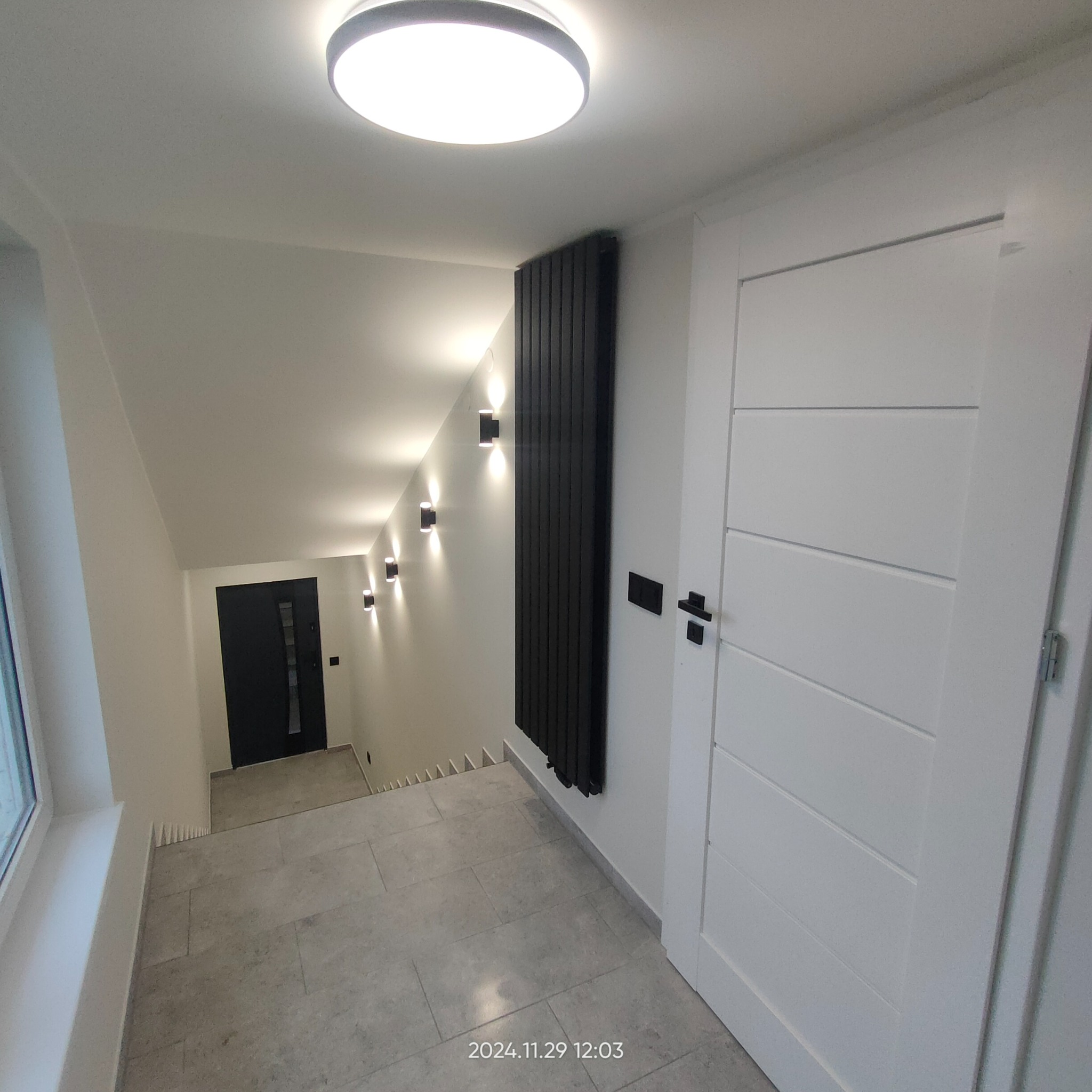 Nowoczesne wnętrze: szare płytki podłogowe, białe ściany z czarnym grzejnikiem panelowym, czarne oświetlenie punktowe, białe drzwi z poziomymi frezami i czarną klamką, widok na schody w dół.