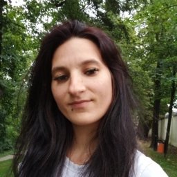 Anna Jeka - Portret kobiety z ciemnymi włosami i piercingiem w wardze, na tle zieleni drzew. Ujęcie z dołu, naturalne światło.