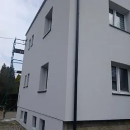 Otynkowana elewacja dwukondygnacyjnego budynku z oknami, rynną i rusztowaniem z boku. Dolna część elewacji z dekoracyjną fakturą. Widok z dołu.