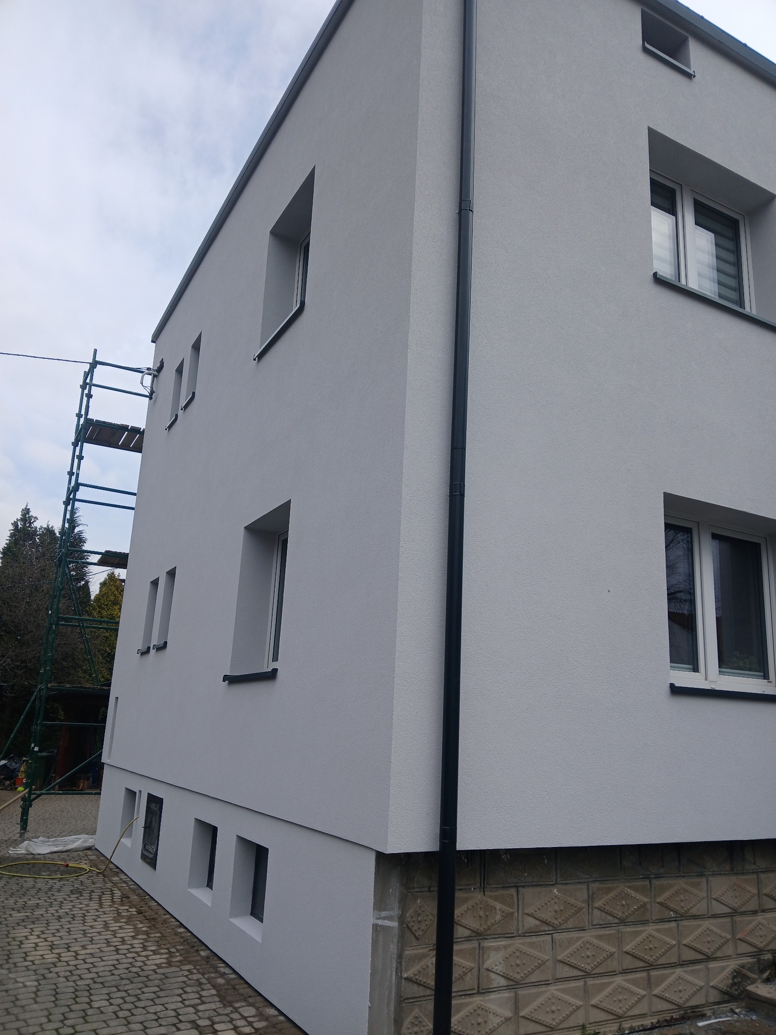 Otynkowana elewacja dwukondygnacyjnego budynku z oknami, rynną i rusztowaniem z boku. Dolna część elewacji z dekoracyjną fakturą. Widok z dołu.