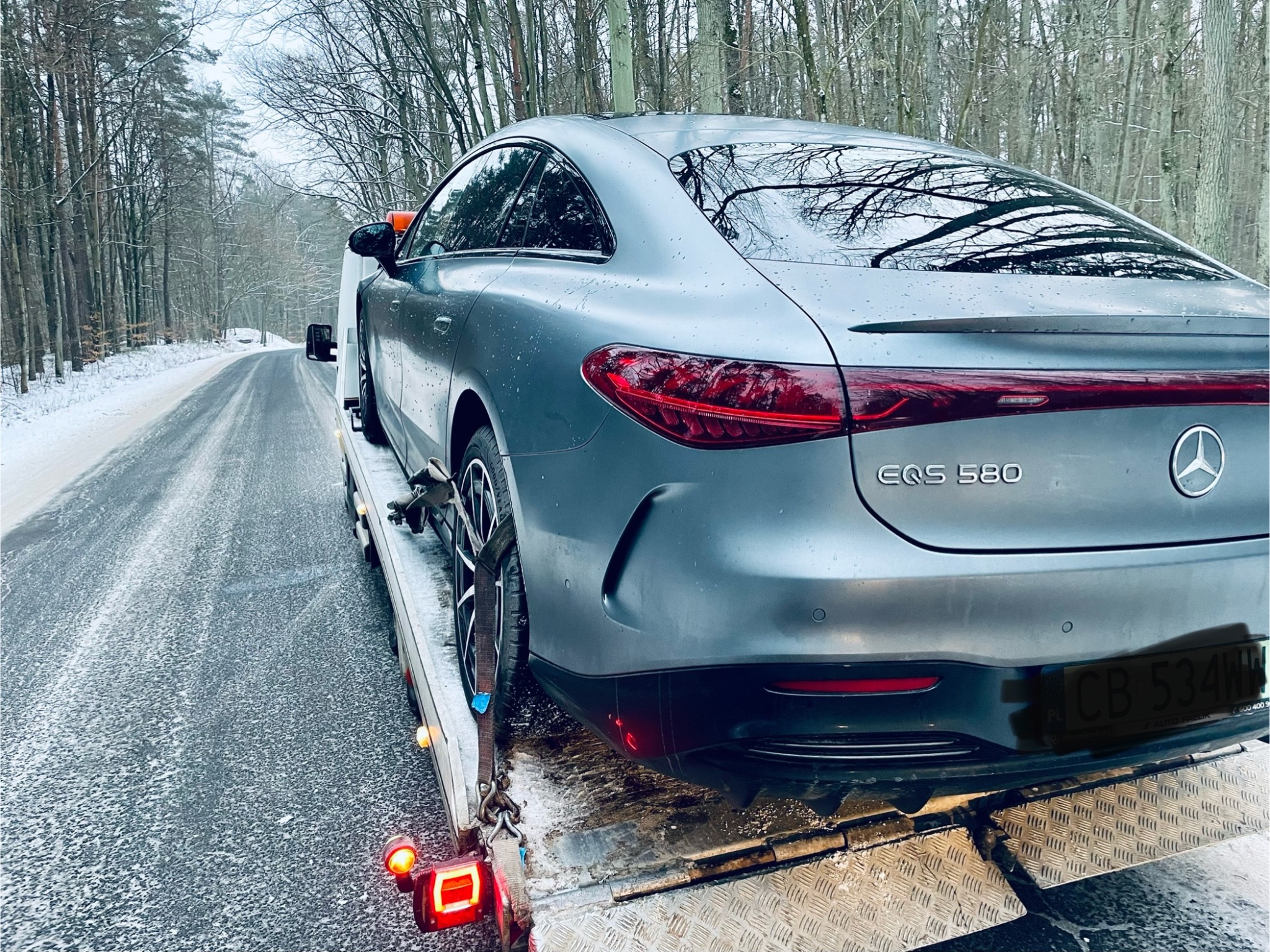 Szary Mercedes EQS 580 zabezpieczony pasami na platformie lawety, widok z tyłu na tle zimowego krajobrazu z ośnieżoną drogą i drzewami.