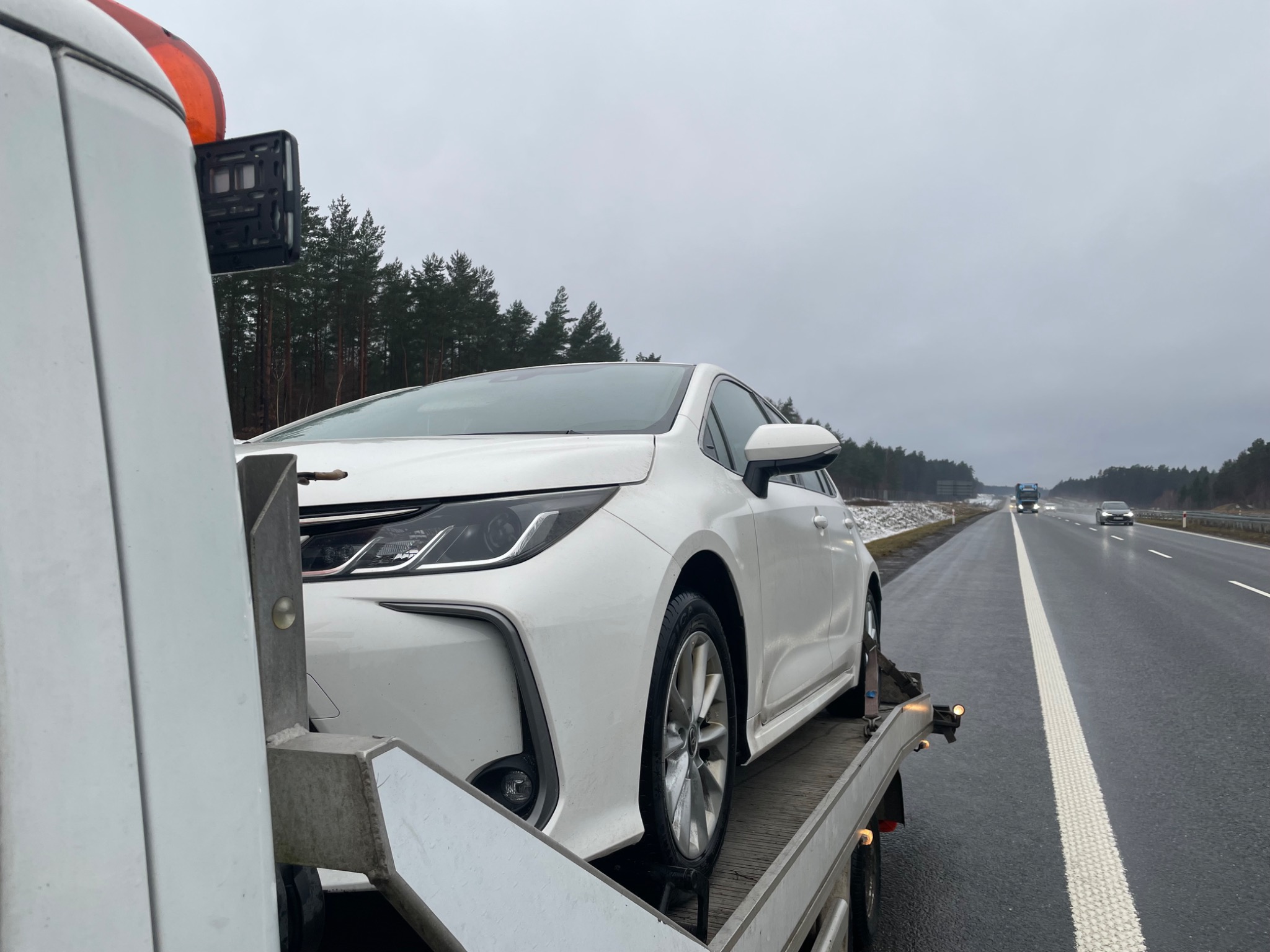 Biała Toyota Corolla na platformie lawety, widok z boku, autostrada w tle, pochmurny dzień.