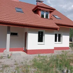 Nowo otynkowany dom jednorodzinny z czerwoną dachówką i ceglaną podmurówką, widok z poziomu gruntu na niedokończony teren.