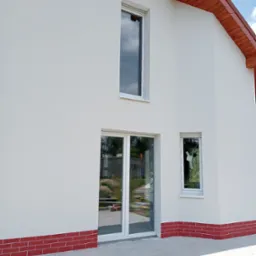 Nowoczesny dom z białą elewacją, czerwonym cokołem z cegły i dużymi oknami, odbijającymi otoczenie.