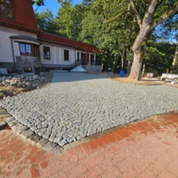 Obszerny plac wyłożony szarą kostką granitową ułożoną w wachlarzowy wzór, obrzeża z ciemniejszej kostki, w tle budynek z czerwonym dachem i drzewa.