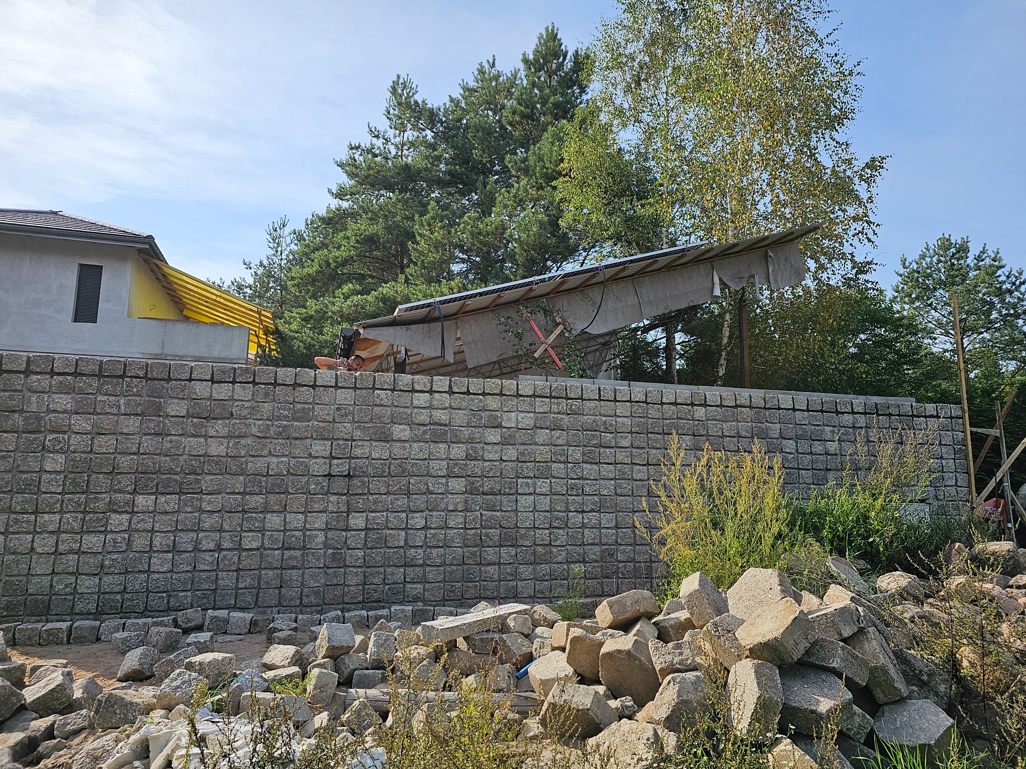 Wysoki murek z kostki granitowej, w tle częściowo widoczny dom i konstrukcja zadaszenia, na pierwszym planie stos kamieni.