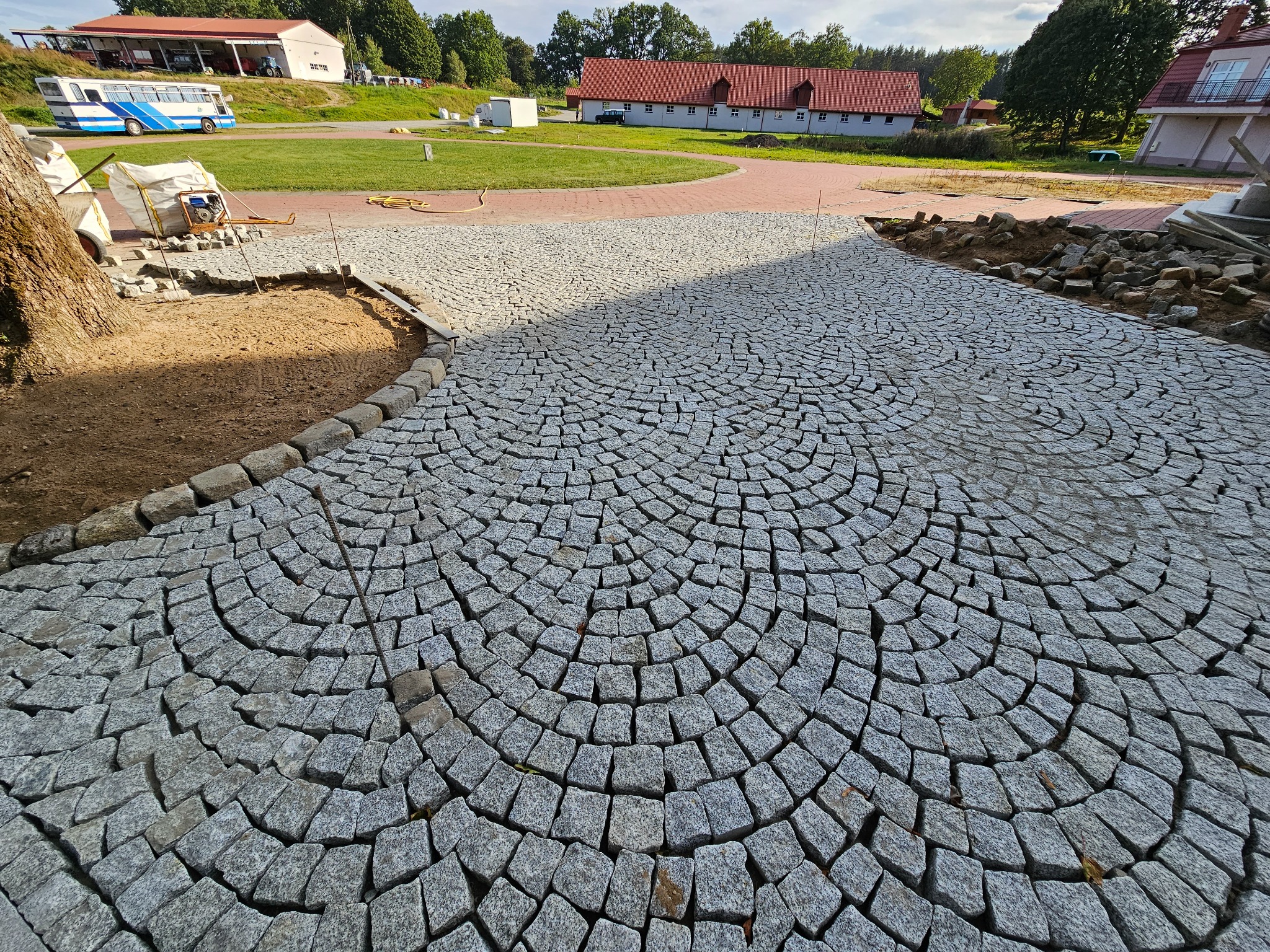 Świeżo ułożona nawierzchnia z kostki granitowej w radialny wzór, z widocznym fragmentem przygotowywanego podłoża i elementami otoczenia: budynki, drzewa.