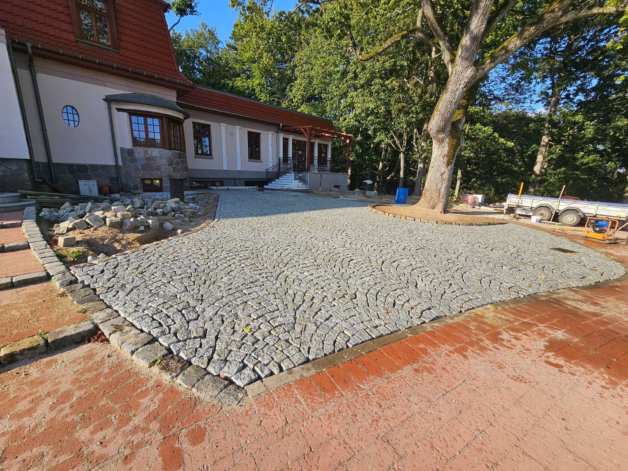Obszerny plac wyłożony szarą kostką granitową ułożoną w wachlarzowy wzór, obrzeża z ciemniejszej kostki, w tle budynek z czerwonym dachem i drzewa.