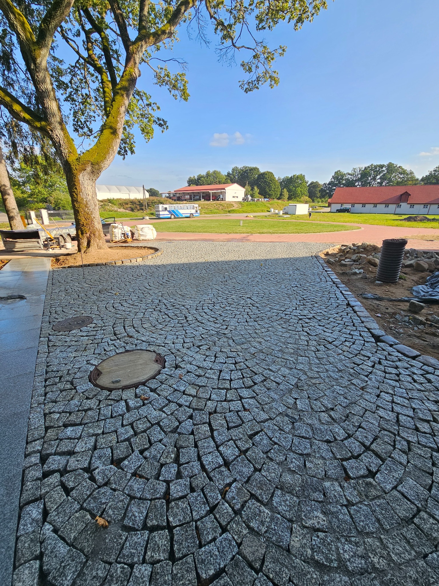 Ułożona kostka granitowa w radialny wzór, z widocznymi studzienkami kanalizacyjnymi, na tle rozległego terenu z budynkami i drzewami w słoneczny dzień.