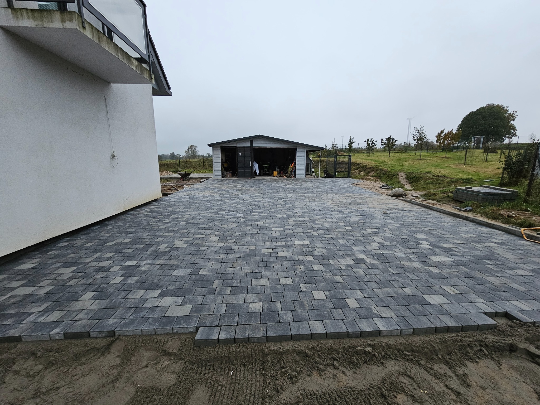 Ułożona szara kostka brukowa na posesji z widocznym garażem blaszanym w tle, częściowo jeszcze niezasypana piaskiem na obrzeżach, pochmurne niebo.