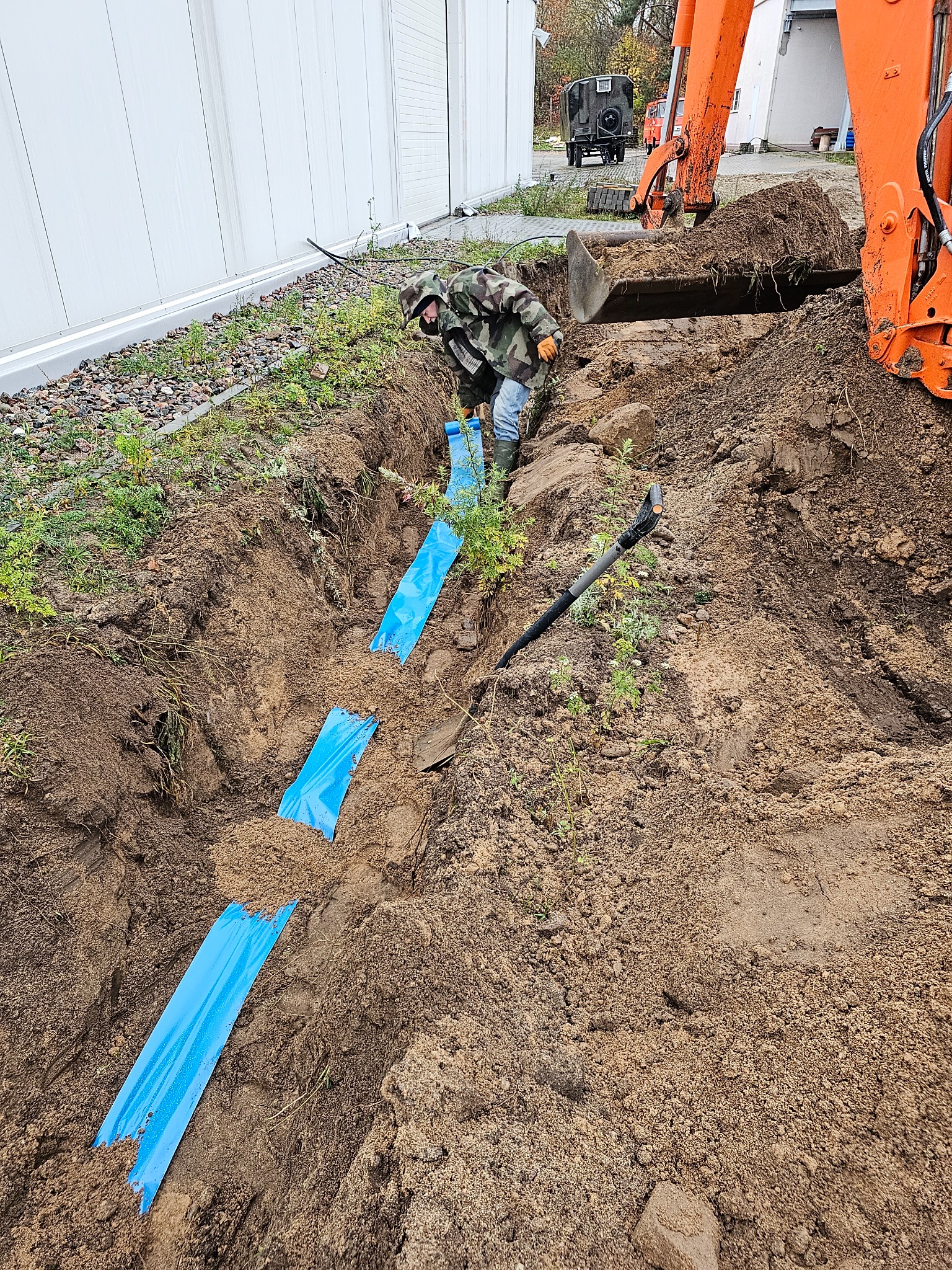 Pracownik w kamuflażu, koparka i wykop oznaczony niebieską folią na tle budynku przemysłowego i pojazdu wojskowego.