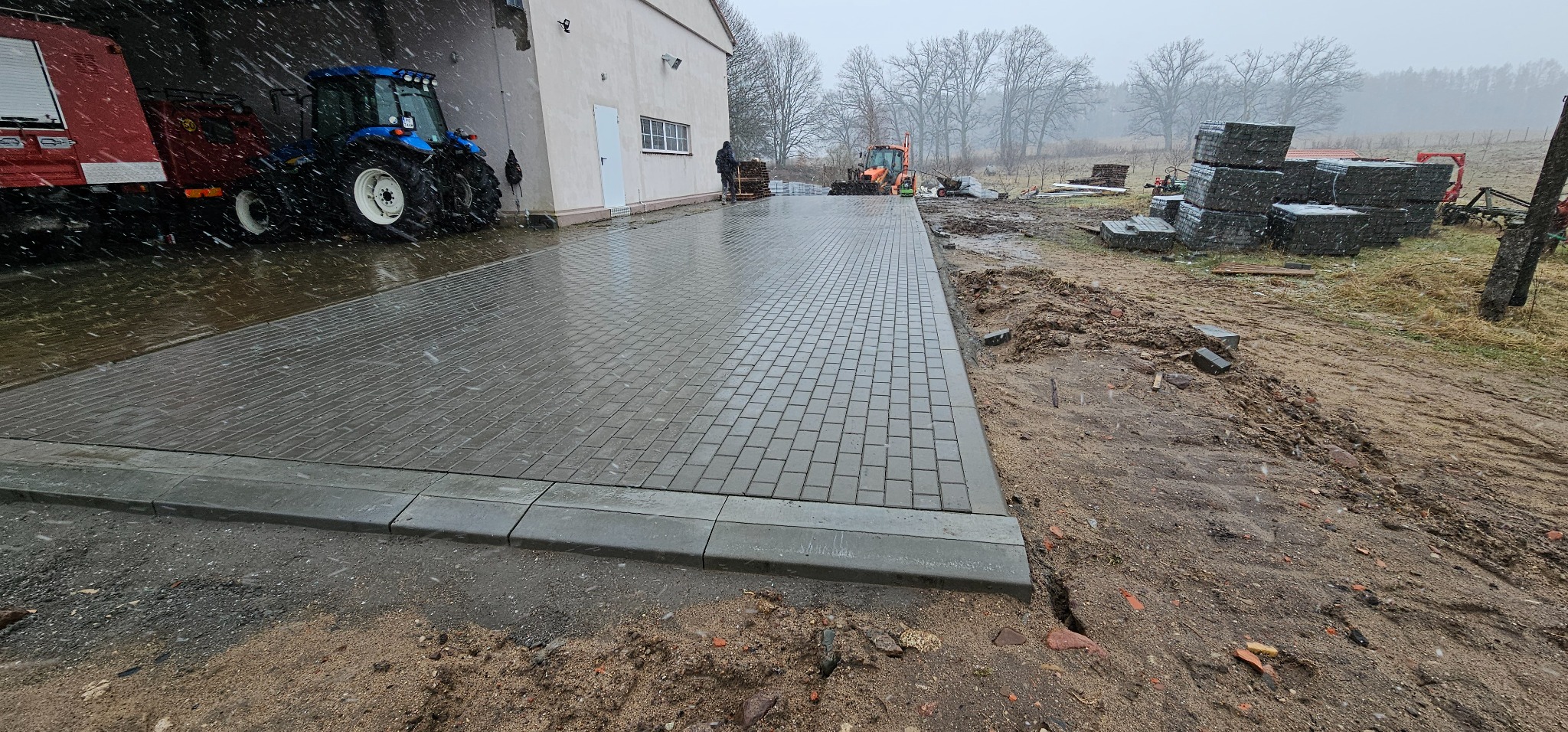Świeżo ułożona, mokra kostka brukowa w szarym odcieniu, otoczona błotnistym terenem i zaparkowanymi maszynami budowlanymi w tle, podczas lekkiego opadu śniegu.