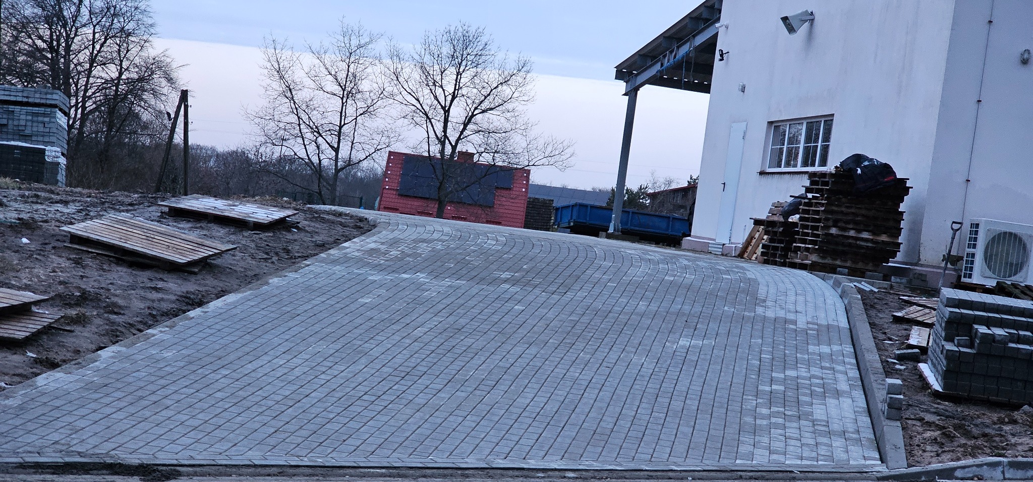 Ułożona szara kostka brukowa tworząca podjazd, widoczne palety z kostką, budynek gospodarczy z klimatyzatorem i drzewa w tle.