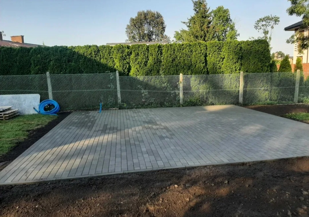Ułożona szara kostka brukowa na przygotowanym podłożu, widoczny fragment trawnika, zwinięty niebieski wąż ogrodowy, siatka ogrodzeniowa i wysokie tuje w tle.