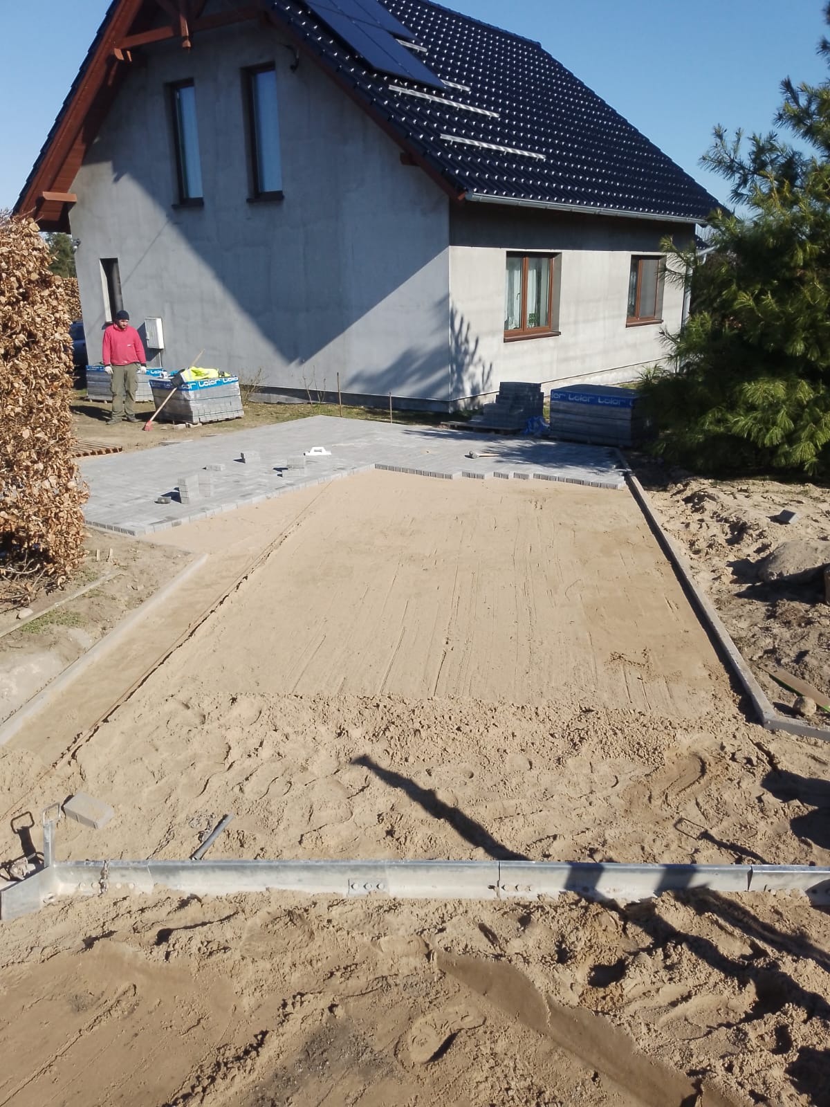 Przygotowanie podłoża z piasku pod ułożenie kostki brukowej, częściowo ułożona kostka przy domu, widoczny pracownik z grabiami oraz palety z kostką w tle.