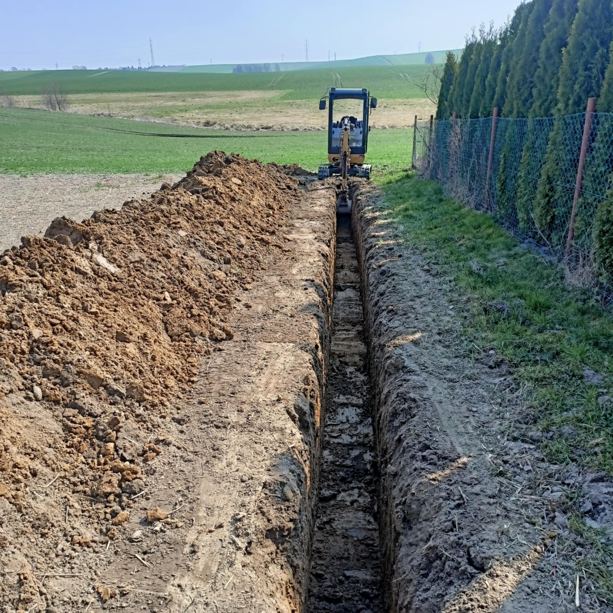 Wąski wykop w ziemi, wykonany przy użyciu małej koparki, z hałdą ziemi po lewej stronie i zielonym polem w tle.