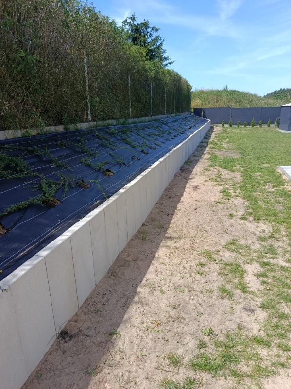 Długi, betonowy murek oporowy z czarną agrowłókniną i młodymi roślinami, zabezpieczający skarpę. Tło: wysokie tuje, trawnik i błękitne niebo.